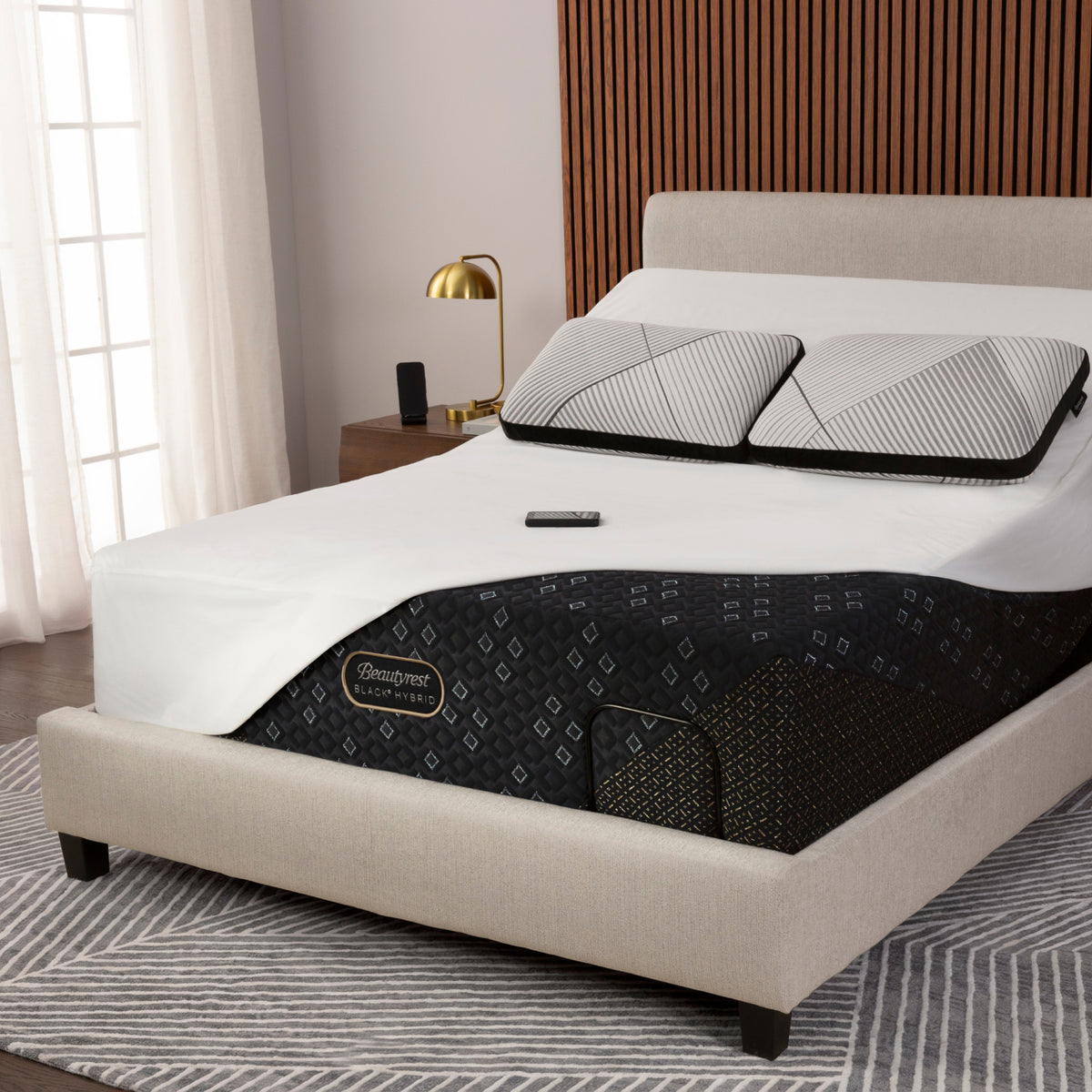 A beautyrest black hybrid mattress dressed in a Beautyrest mattress protector and two striped pillows, with a black mattress or mattress topper showing the Quimper brand label. The bed is set in a modern bedroom with a beige upholstered frame, a wooden slat headboard, a gold bedside lamp, and a gray patterned rug.