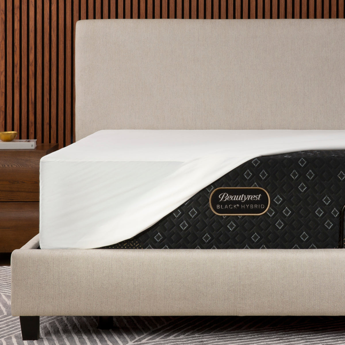 Beautyrest Black Hybrid mattress shown on a bed frame with a Beautyrest mattress protector, set against a modern wood slat wall background.
