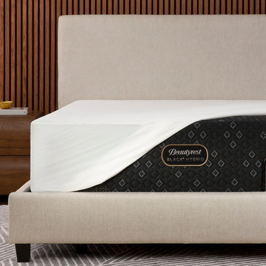 Beautyrest Black Hybrid mattress shown on a bed frame with a Beautyrest mattress protector, set against a modern wood slat wall background.