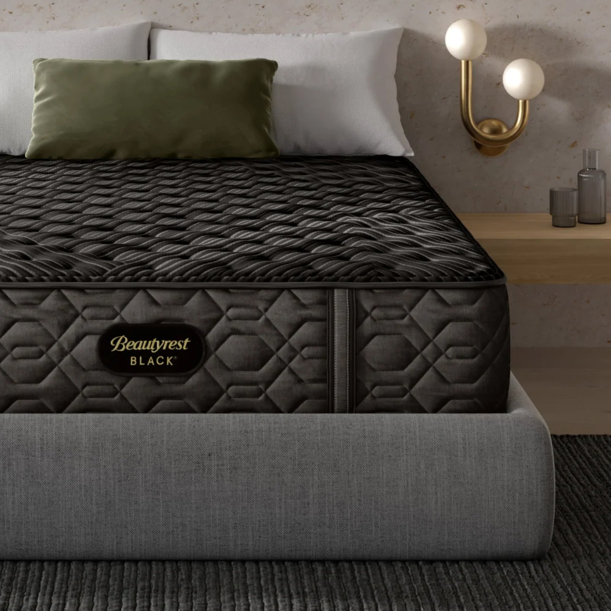 A Beautyrest Black® Innerspring mattress on a grey upholstered bed frame, featuring a quilted black surface with a textured pattern, paired with white and green pillows.